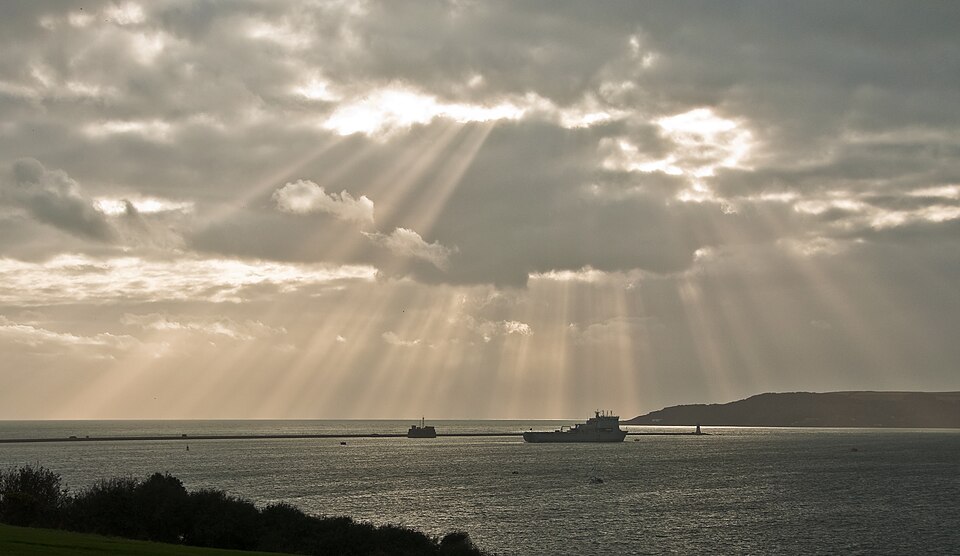 Sunbeams shining through dramatic clouds over Plymouth Sound, with ships on the water and distant coastline beneath the evening light.