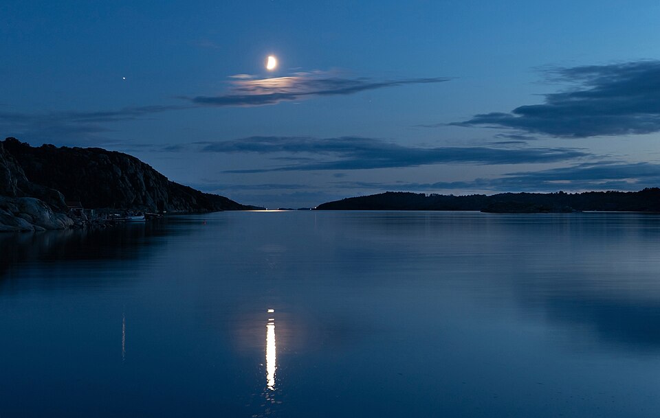 Waxing half moon shining over calm blue water at Brofjorden, with moonlight reflection, rocky shoreline, and peaceful evening sky.
